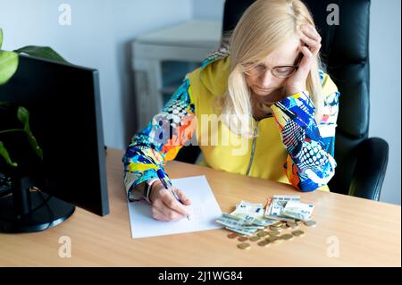 A middle-aged thoughtful woman counting euro money and takes notes at a desk in the home office Stock Photo