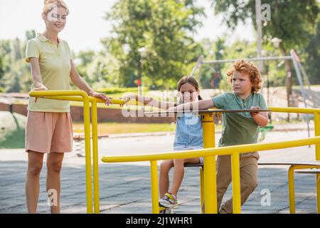 cheerful woman standing near kids on carousel in park Stock Photo