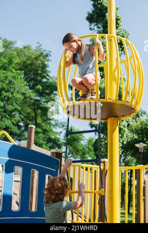 boy climbing on rope ladder under sister on high tower in amusement park Stock Photo