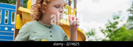 curly boy blowing soap bubbles while spending time outdoors, banner Stock Photo