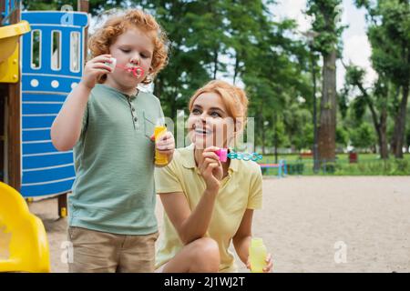 redhead boy blowing soap bubbles near cheerful mother on playground Stock Photo