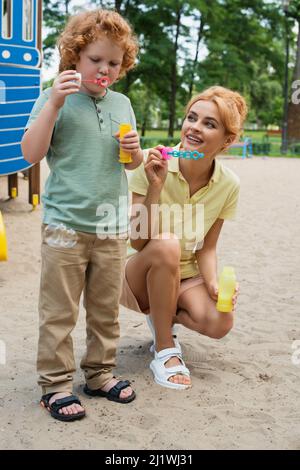 full length view of happy woman near son blowing soap bubble on playground Stock Photo