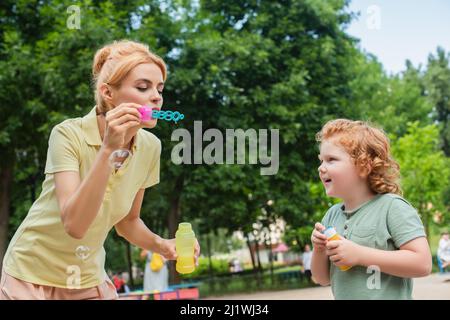 redhead woman blowing soap bubbles near excited son on playground Stock Photo