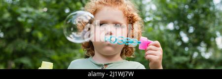 curly child blowing soap bubble outdoors, banner Stock Photo