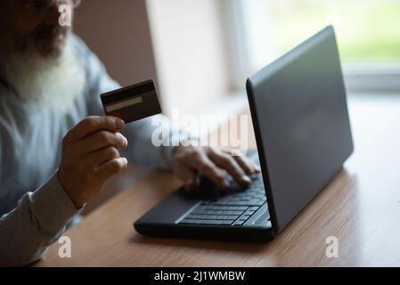 Portrait of bearded senior elderly retired man sitting at the  laptop computer and  holding credit bank card Stock Photo