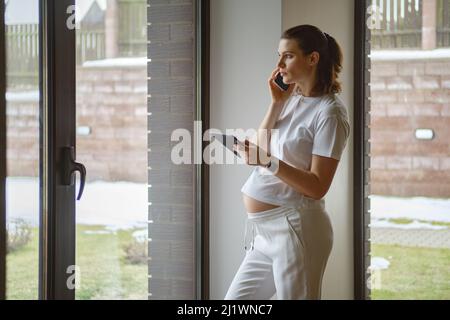 Attractive woman does business from home during pregnancy, making phone call with tablet in hand Stock Photo