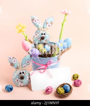 Cute children Easter card with small blue bucket, nests, colored quail eggs, decorative eggs, knitted color flowers, textile bunnies, empty note on pa Stock Photo