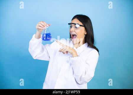 Young brunette woman wearing scientist uniform holding test tube over isolated blue background amazed and pointing with hand and finger Stock Photo