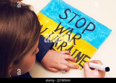 Little Ukrainian girl draws a picture with the flag of Ukraine and the text STOP WAR IN UKRAINE. Russian invasion of Ukraine. Stock Photo
