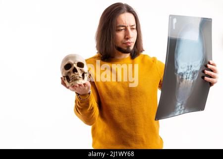 Intrigued young caucasian bearded guy comparing artificial skull model to big skull x-ray over white background. Health concept. . High quality photo Stock Photo