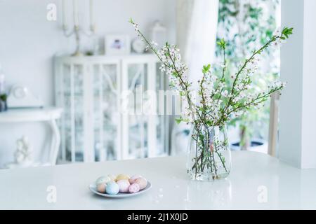 Blooming tree branches in the vase and colored easter eggs on the plate on white kitchen table with classic style interior background. Easter celebrat Stock Photo