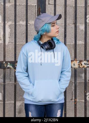 Teenager wearing light blue oversize hoodie and baseball cap looking right. Blue haired teen girl with wireless headphones stays outdoor near a gate. Stock Photo