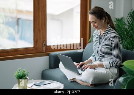 Asian young woman working from home office sit on couch hold paper invoice check information online Stock Photo