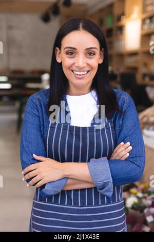 Portrait of smiling hispanic young female barista with arms crossed wearing apron in coffee shop. unaltered, cafe culture, people and occupation conce Stock Photo