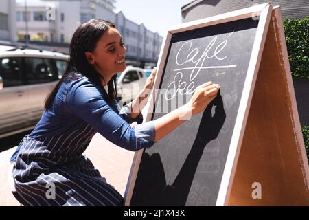 Smiling young caucasian female coffee shop employee writing on chalkboard at sidewalk. unaltered, cafe culture, people and occupation concept. Stock Photo