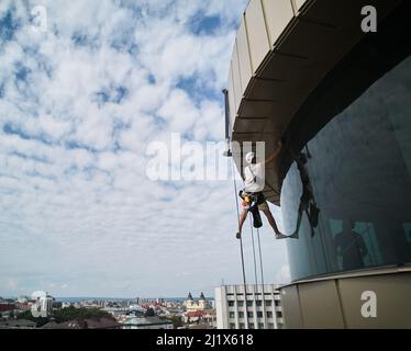 Industrial mountaineering worker cleaning glass window of skyscraper. Male cleaner using safety lifting equipment while washing window of high-rise building. Stock Photo