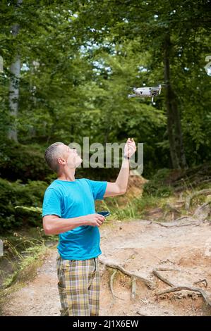 Man operating drone using remote controller. Man using drone for photos and video making while standing in green forest. Stock Photo