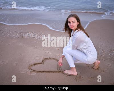 A young woman in white on the beach drawing a heart in the sand and looking at the camera. Stock Photo