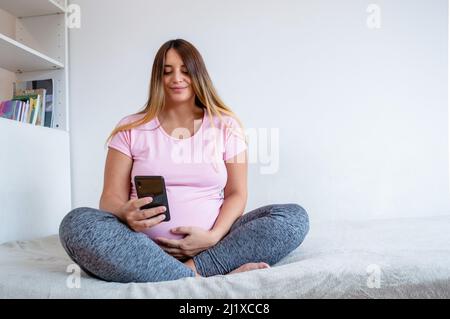 beautiful young pregnant Caucasian Venezuelan woman sitting on her bed in her apartment using the phone, messaging her husband, surfing the internet. Stock Photo