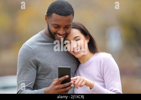 Front view of a happy interracial couple checking smart phone outdoors in a park