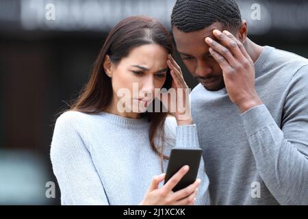 Worried interracial couple checking smart phone standing in the street Stock Photo