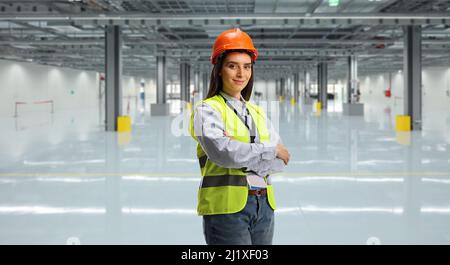 Young female engineer wearing reflective vest and helmet and standing in an empty factory hall Stock Photo