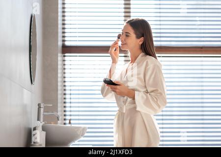 Young Female In White Silk Robe Applying Makeup Near Mirror In Bathroom Stock Photo