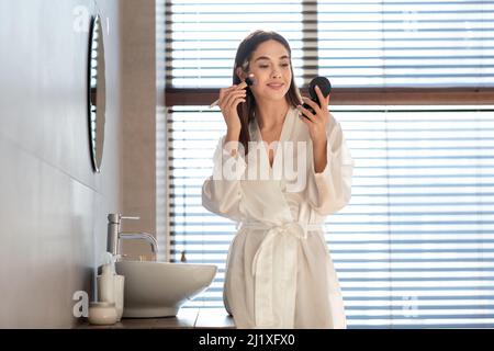 Getting Ready. Attractive Woman In White Silk Robe Applying Blush On Cheeks Stock Photo