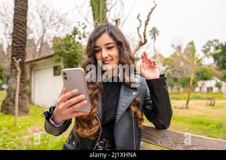 Happy young woman sitting on a park bench holding phone looking at screen waving hand video calling distance friend online mobile chat app. Smartphone Stock Photo