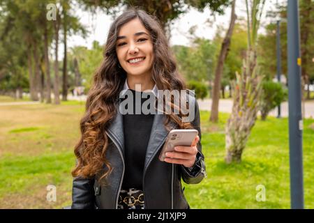 Smiling young lady, business woman posing for the camera and holding her phone, she is spending her time in an outdoor park and smiling, wavy hair and Stock Photo