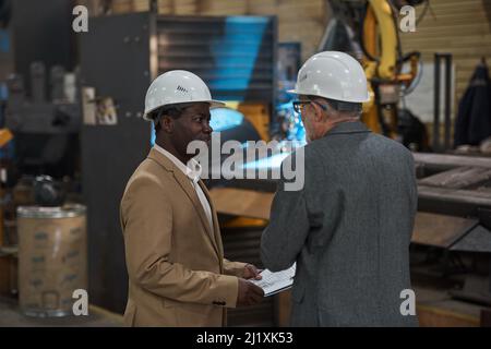 Rear view of two engineers in suit and hardhats discussing the work of machine in the factory Stock Photo