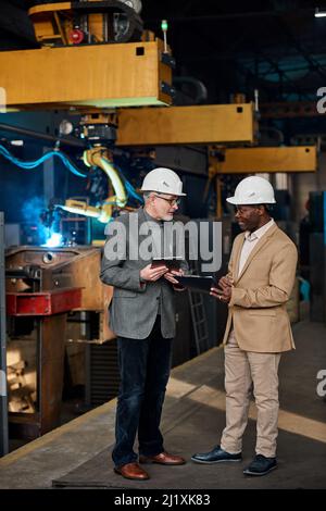 Multiethnic business people discussing the work of production in team standing in factory with big machines in the background Stock Photo