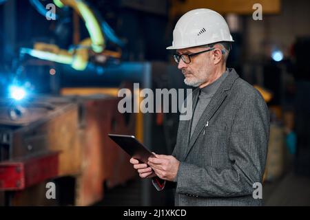 Serious elder engineer in work helmet using digital tablet to control the work on factory Stock Photo
