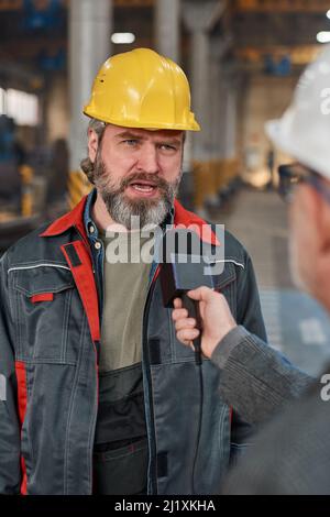 Mature male worker in hardhat and uniform talking in microphone while giving interview about machine factory to reporter Stock Photo
