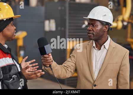 African journalist in work helmet holding microphone and talking to female worker in plant Stock Photo