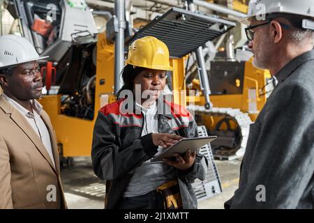 African female worker in uniform using tablet pc and presenting her report to engineers while they standing in warehouse with machines in the backgrou Stock Photo