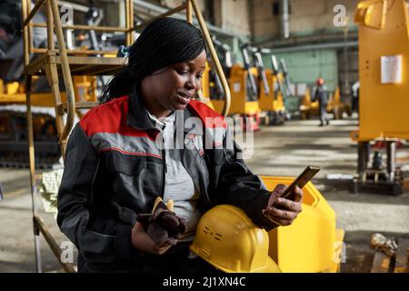 African female worker in uniform reading a message on mobile phone while sitting in warehouse during her break Stock Photo