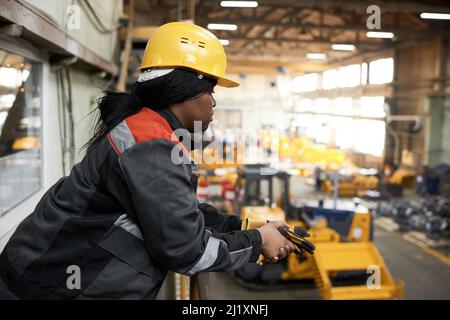 African female worker in uniform and hardhat standing on balcony and watching at empty warehouse with machines Stock Photo