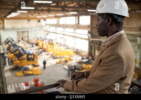 Serious African engineer in work helmet standing on balcony and watching for the work of machines in factory Stock Photo