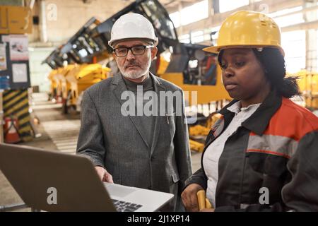 Senior engineer in work helmet using laptop to present new machines to worker who looking at monitor standing near Stock Photo