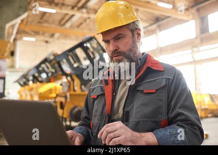 Mature bearded foreman in uniform working online using laptop to control the equipment in warehouse Stock Photo
