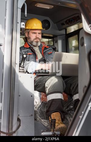 Mature engineer in uniform sitting in cabin and controlling the work of machine with laptop Stock Photo