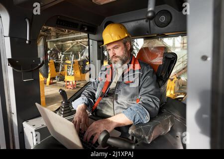 Mature operator in uniform and work helmet sitting in cabin of construction machine and controlling the work with laptop Stock Photo
