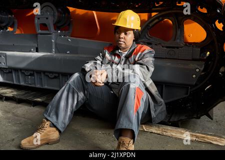 African female worker in uniform and hardhat sitting on the floor leaning on tractor and resting after work Stock Photo