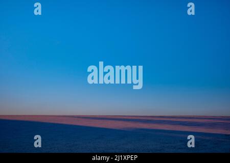 AT the end of the day as the sunsets a  perfect blue sky over a filed covered in snow with deep sahdow and oink streaks of light, Stock Photo