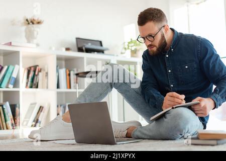 Millennial Guy Writing Taking Notes Looking At Laptop At Home Stock Photo