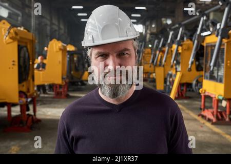 Portrait of mature bearded worker in work helmet looking at camera while standing in warehouse Stock Photo