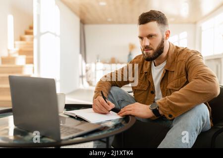 Side View Of Man Taking Notes And Using Laptop Indoor Stock Photo