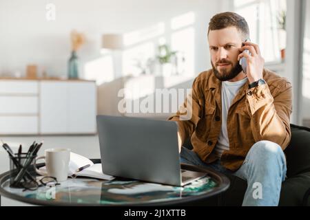 Serious Male Freelancer Talking On Phone Using Laptop At Home Stock Photo