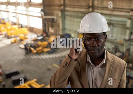 African engineer in work helmet looking very serious while talking on mobile phone with partner standing in plant Stock Photo